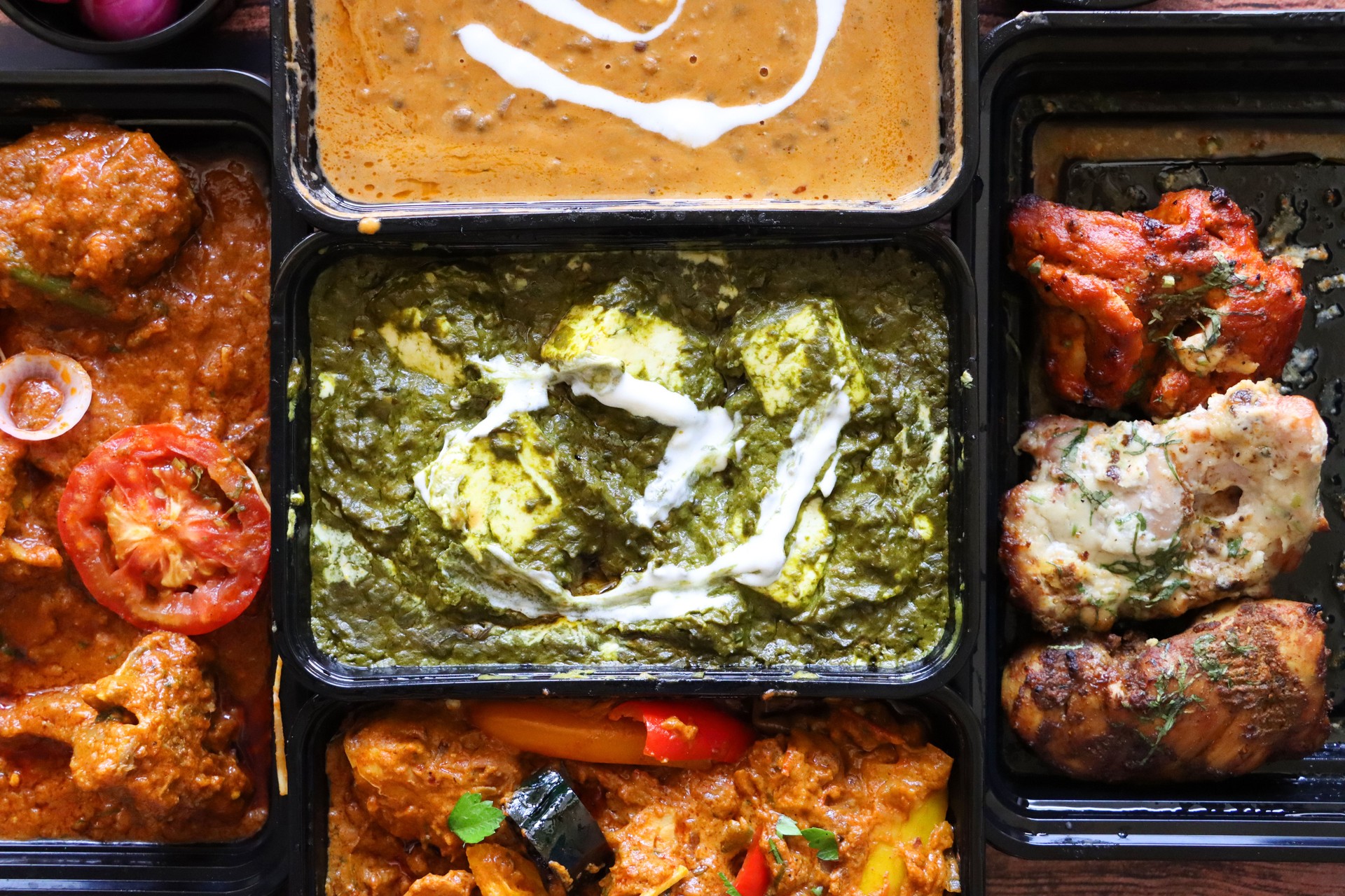 Image of selection Indian takeaway dishes in black plastic, single-use food containers, recipes including, Kadai Chicken, Chicken Bhuna, Palak paneer, Murgh malai tikka (grilled chicken supreme) and Dal makhani (black lentil curry), elevated view